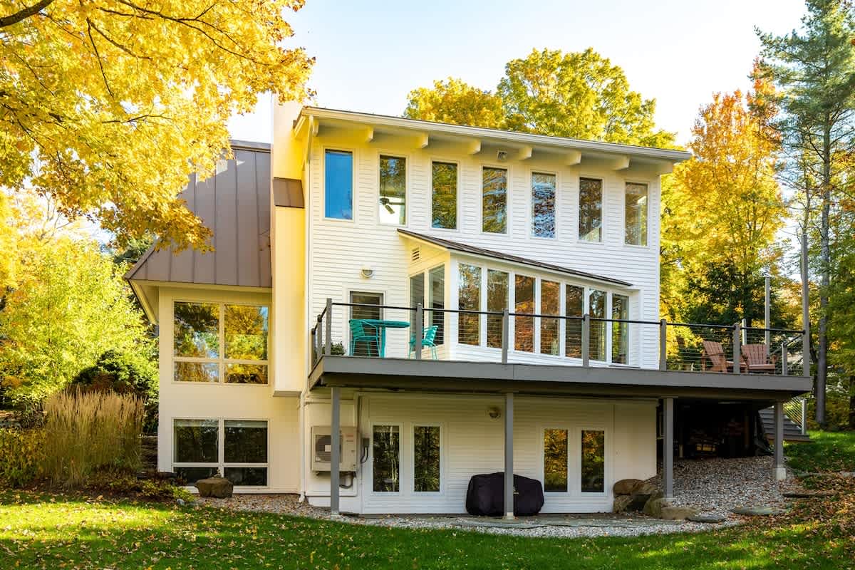 Multi-level white house with large windows and a spacious deck featuring outdoor seating, surrounded by bright yellow autumn trees, showcasing a walk-out basement and a green lawn.