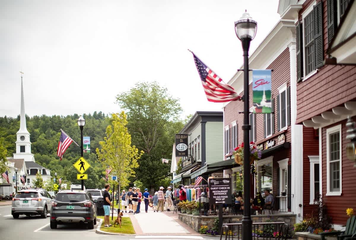 A lively small-town street features charming storefronts, parked cars, and pedestrians strolling along the sidewalk. American flags adorn lampposts, and a white church steeple is visible in the distance, capturing a classic American town atmosphere.