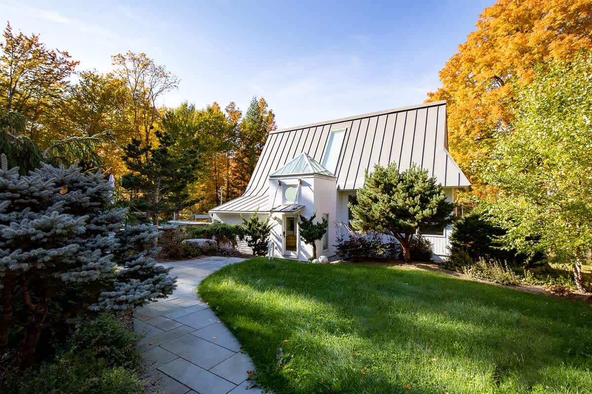Modern white house with a unique curved metal roof and a prominent skylight, surrounded by vibrant autumn foliage and a manicured lawn with a stone pathway leading to the entrance.