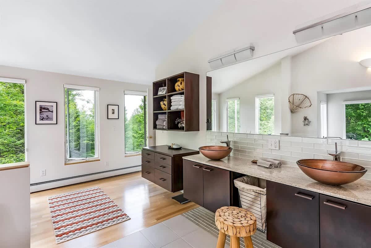 Bright and airy bathroom with light tile flooring and a double vanity featuring copper vessel sinks and dark wood cabinetry. Large windows offer natural light and views, enhancing the modern aesthetic.