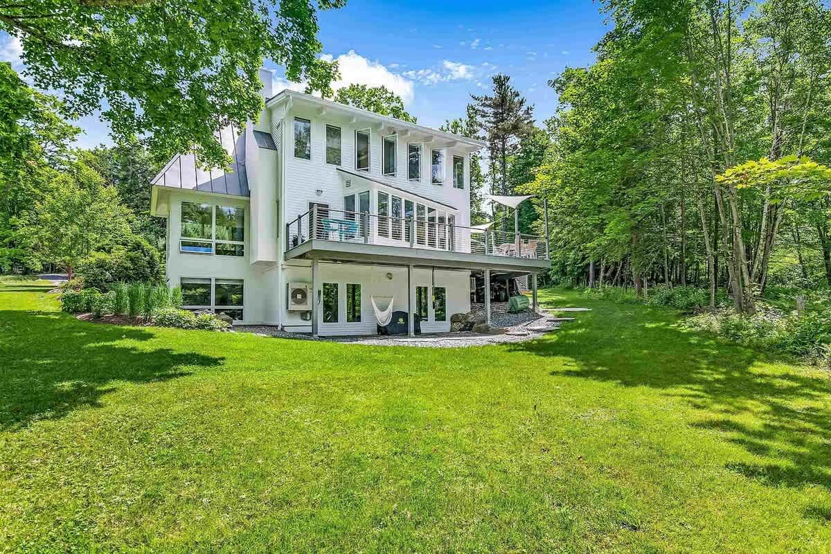 Expansive back view of the white multi-level house with a large deck, glass railings, and a walk-out basement, overlooking a spacious green lawn surrounded by mature trees.