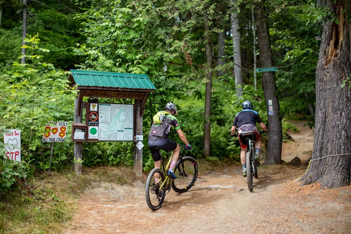 Two mountain bikers are on a dirt trail in a lush green forest, passing a wooden sign with a map for 'Gary Hill Forest'. The biker in front is doing a wheelie, showcasing an active outdoor scene.