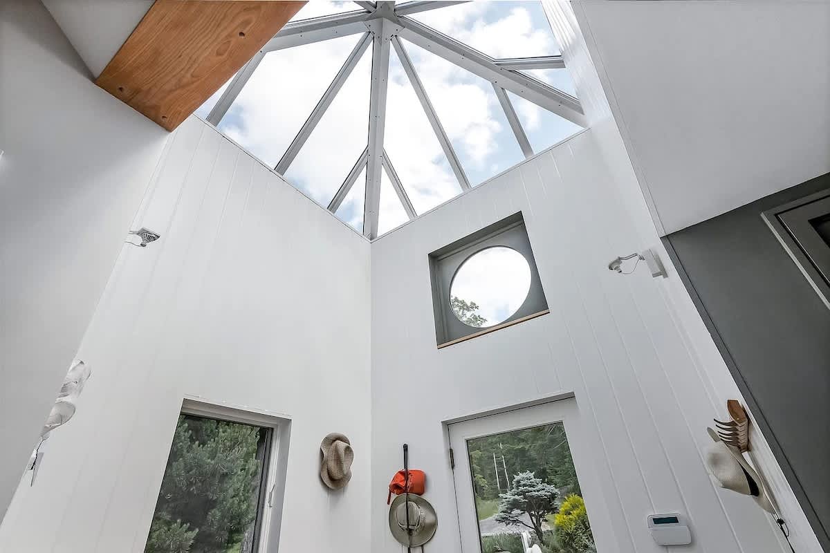 Bright entryway with a high ceiling and a large geometric skylight, featuring white paneled walls, a round window, and a glass-paned door leading outside.