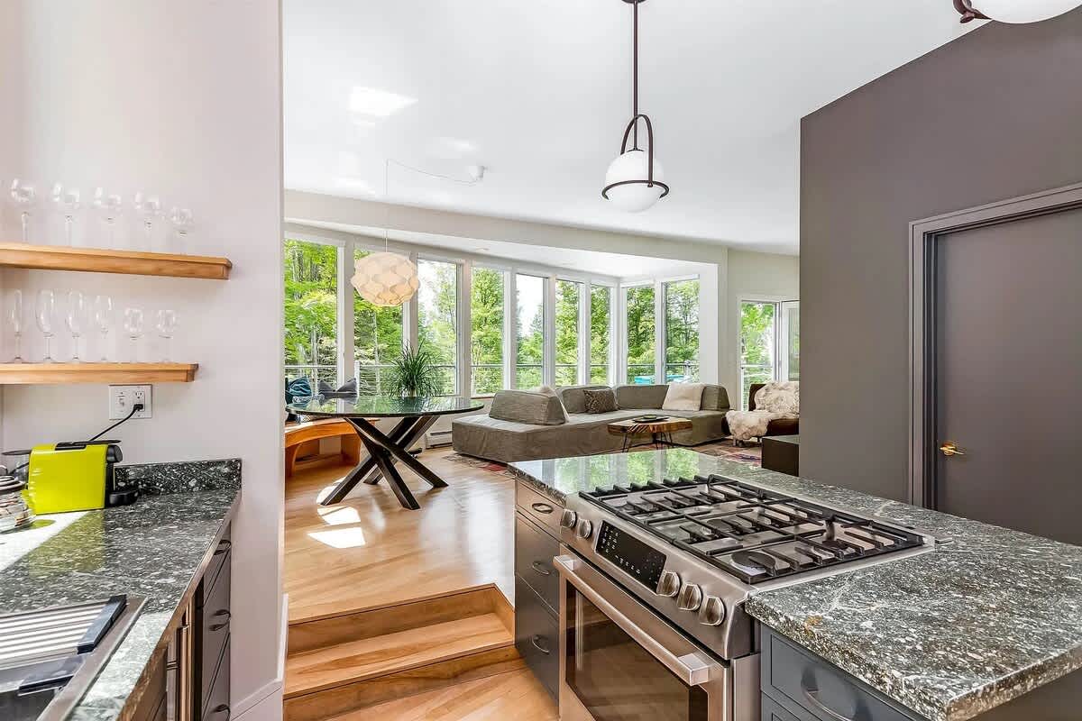 View from the kitchen into an open-concept dining and living area, featuring a large granite island with a gas range. The space is brightly lit by natural light from numerous windows, showcasing hardwood floors and modern decor.