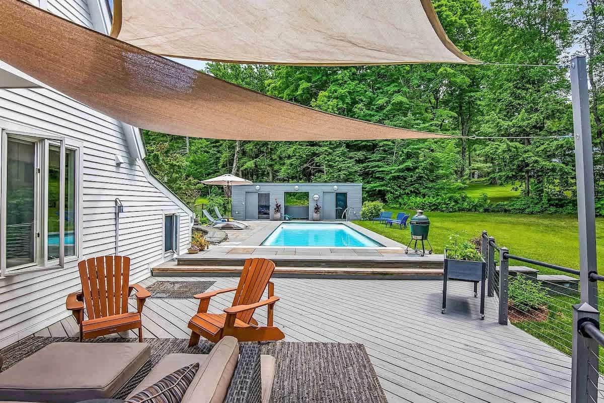 A wide shot of the outdoor deck with a large shade sail, comfortable seating, and a BBQ grill, leading down to a rectangular swimming pool surrounded by a stone patio and green lawn.