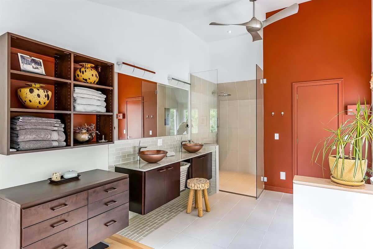 Spacious modern bathroom with a double vanity featuring copper vessel sinks and dark wood cabinets, set against an orange accent wall. It includes a large glass-enclosed shower and a ceiling fan.