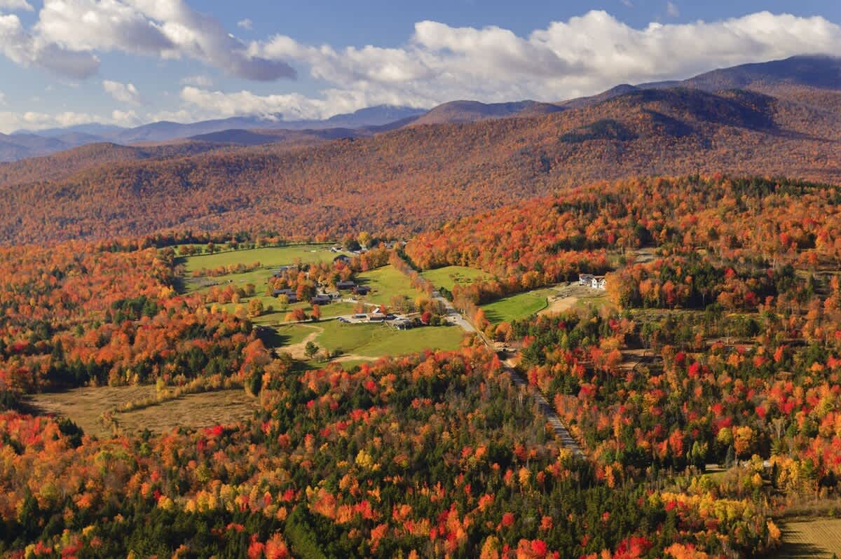 An aerial view showcases a valley bursting with vibrant autumn colors, featuring a mix of red, orange, and yellow trees. Scattered houses and green fields are nestled among the colorful forest, with mountains in the distance.