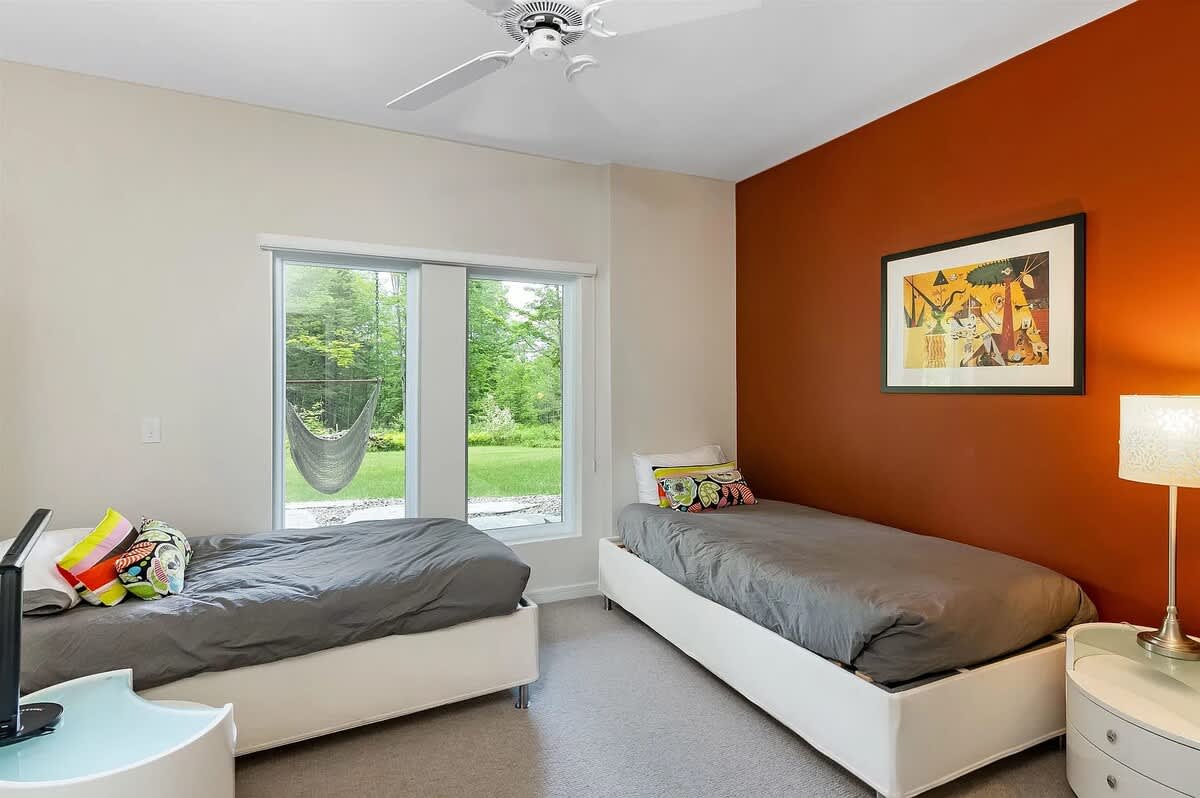A bright bedroom with two twin beds, one against a warm rust-colored accent wall adorned with framed artwork, and large windows overlooking a green outdoor space with a hammock.