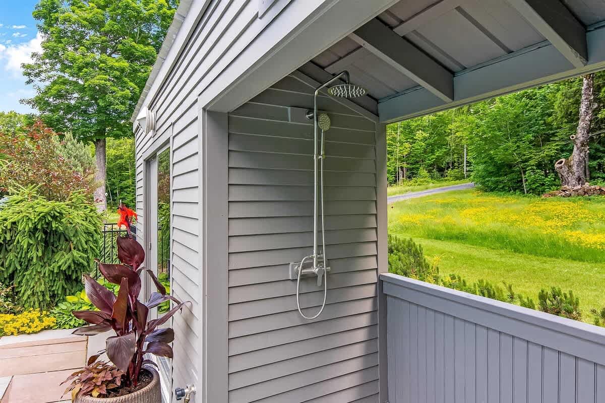 An outdoor shower with a modern rain showerhead mounted on the side of a gray-sided building, surrounded by lush greenery and a potted plant, near the pool area.