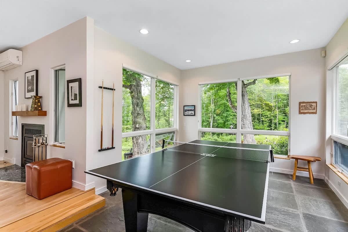 Bright game room with large windows offering views of trees, featuring a black ping pong table, slate tile flooring, and a small seating area with a fireplace in the corner.