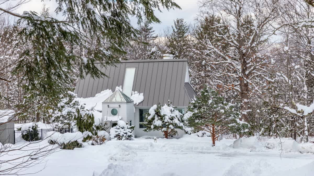 White house with a steep, snow-covered metal roof and a unique turret-like entrance, surrounded by snow-laden evergreen and deciduous trees under a winter sky.