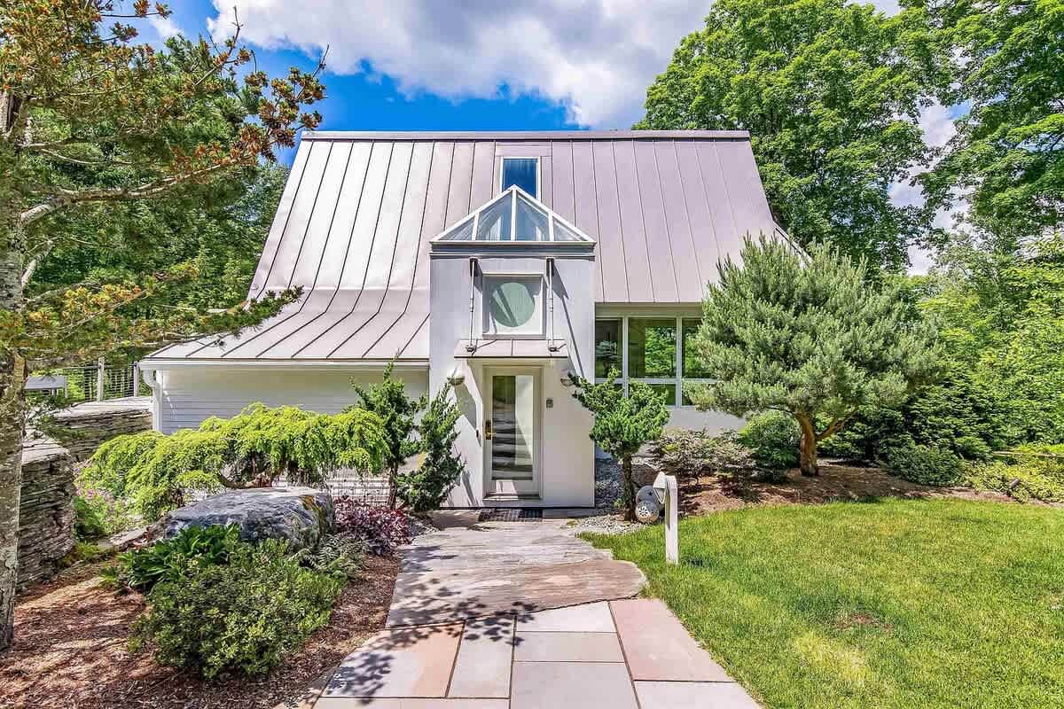 The front exterior of a modern house features a distinctive light gray standing seam metal roof and white siding. A stone pathway leads to the front entrance, which is highlighted by a glass-enclosed structure and a circular window above the door, surrounded by lush green landscaping and mature trees.