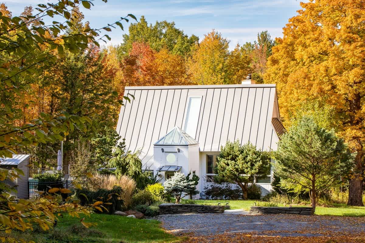 Contemporary white house with a distinctive curved metal roof and a central skylight, nestled among colorful autumn trees with a gravel driveway and landscaped garden beds.