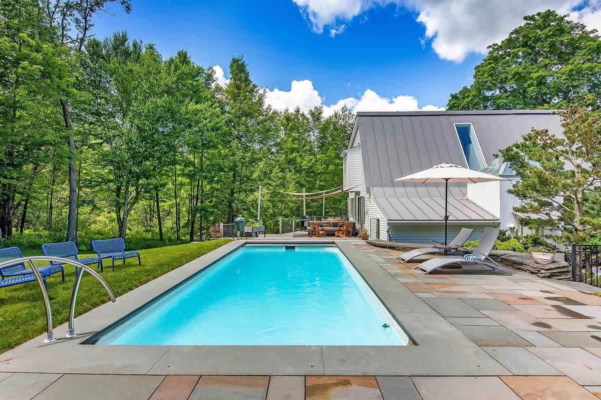 Rectangular swimming pool with light blue water, surrounded by a concrete patio and green grass, with lounge chairs and an umbrella, and a modern house with a grey metal roof in the background.