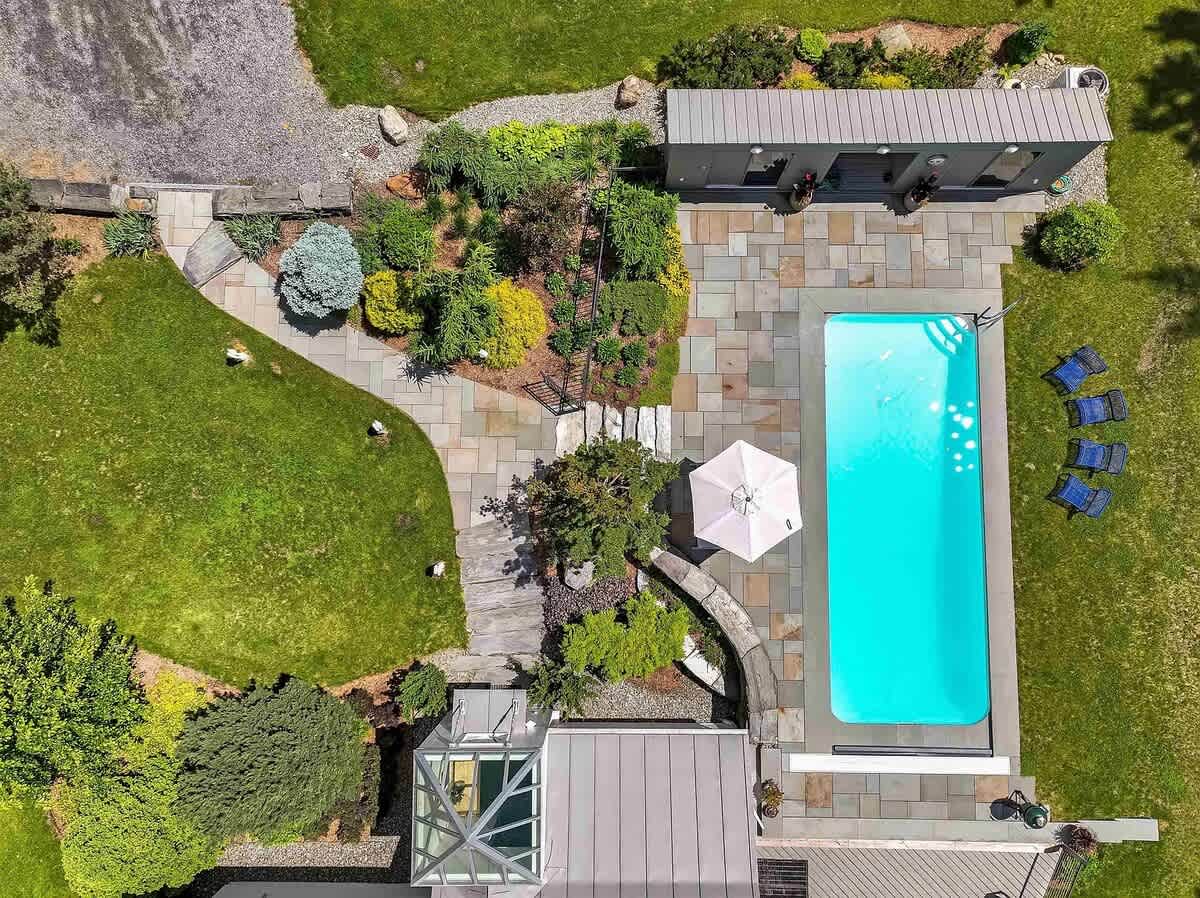 An aerial view showcasing a rectangular swimming pool surrounded by a stone patio and lush green lawn, with a modern pool house and landscaped garden areas.