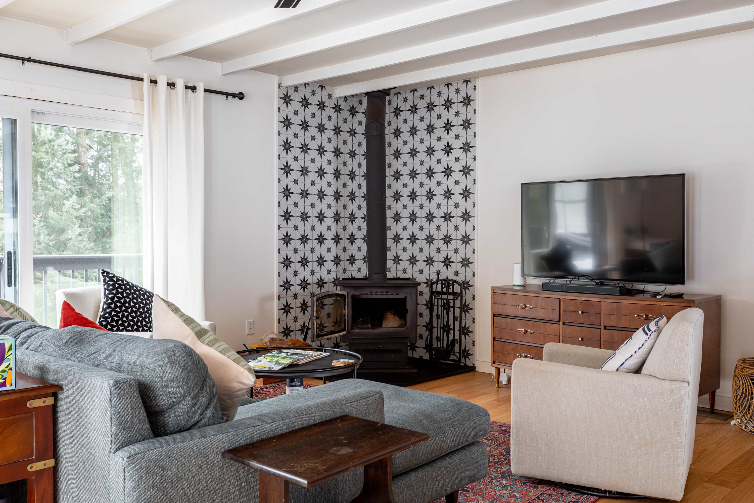 Cozy living room featuring a grey sectional sofa with colorful throw pillows, a black round coffee table, and a woodstove against a star-patterned accent wall. Sliding glass doors offer views of the outdoors.