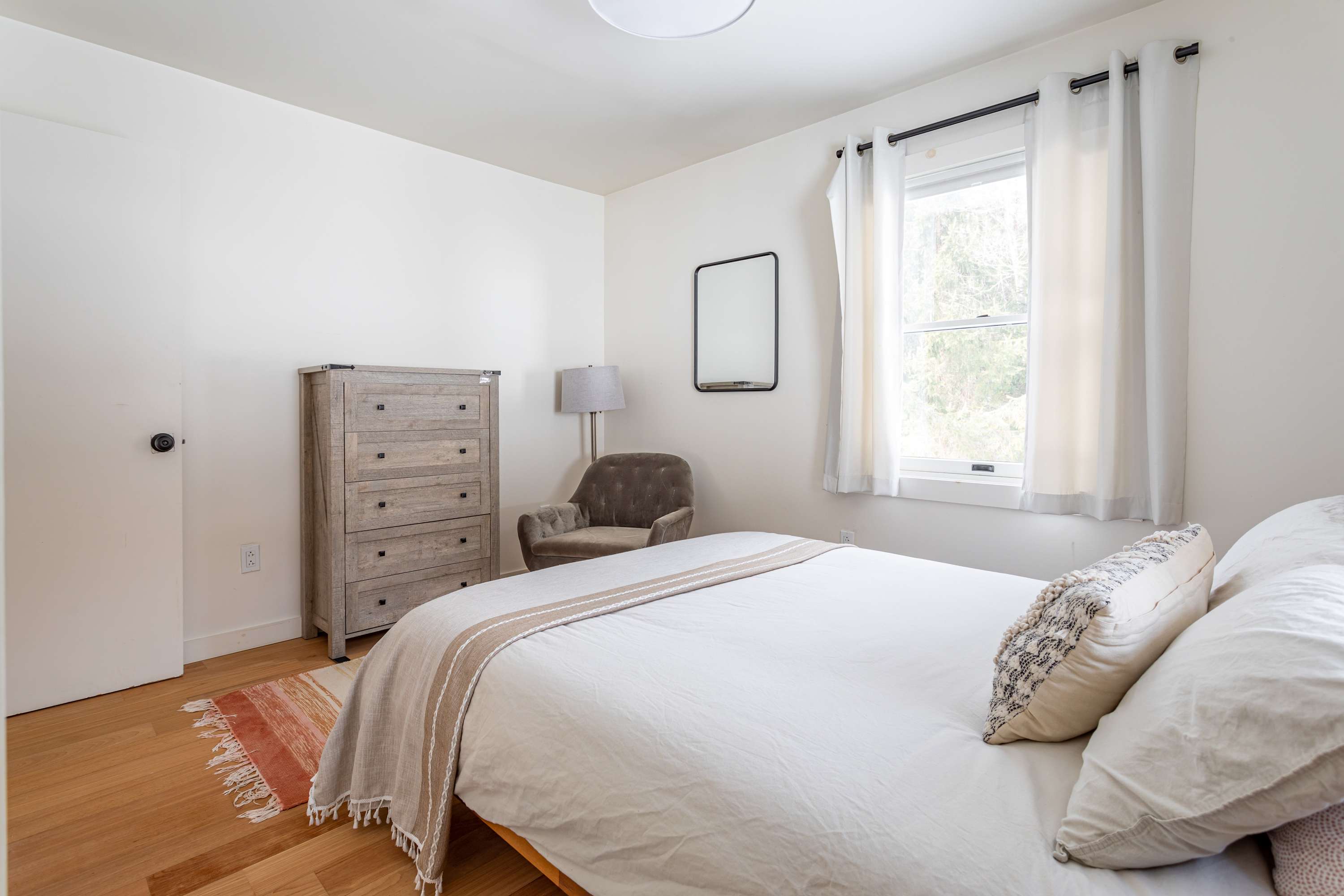 Serene bedroom featuring a queen-sized bed with a wooden headboard, a rustic wooden dresser, and a plush armchair in the corner. A large window provides natural light, and hardwood floors complete the look.