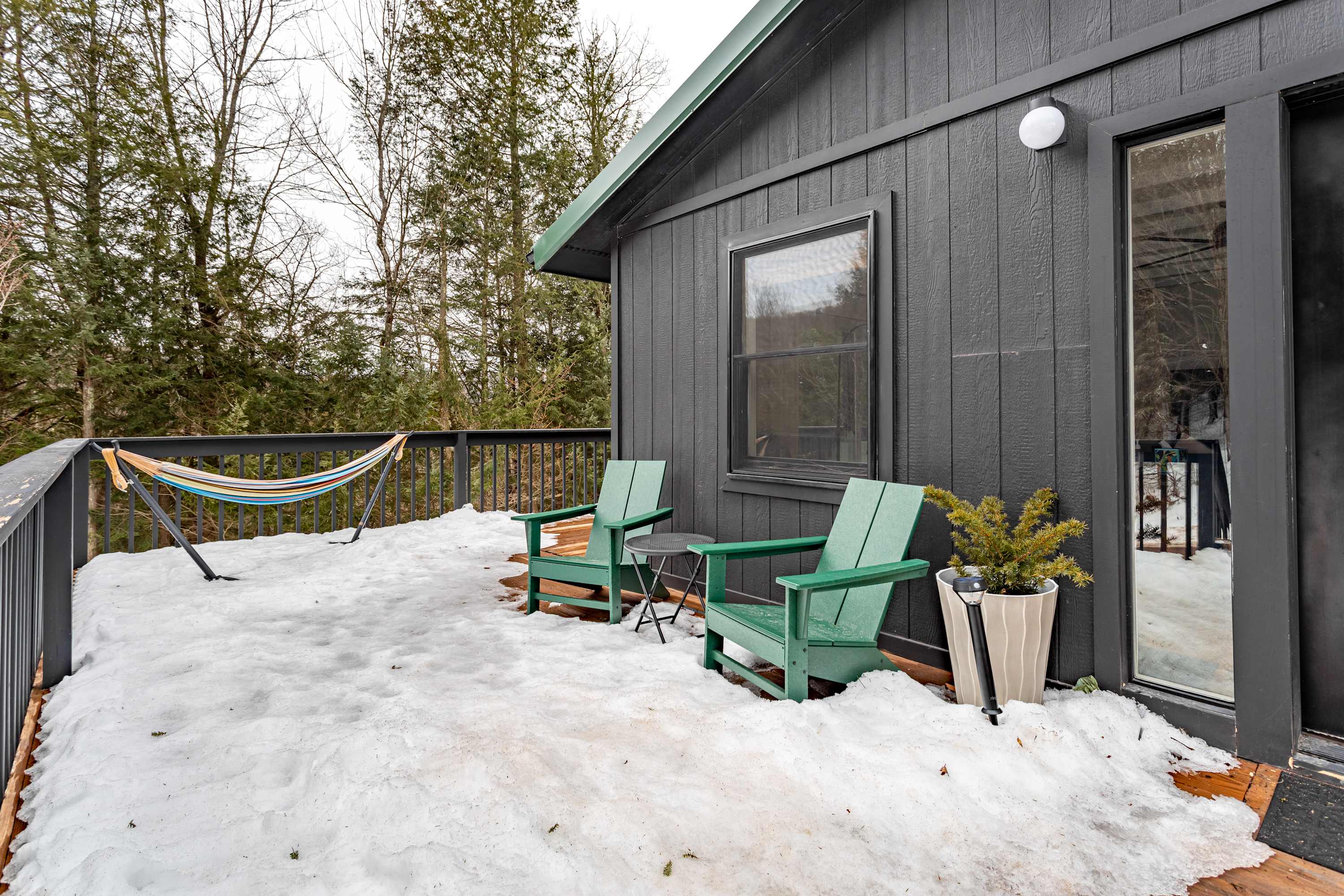 A wooden deck, partially covered in snow, features two green Adirondack chairs, a small side table, and a colorful striped hammock. The dark gray modern cottage exterior with a green roof trim is visible, surrounded by tall evergreen trees in the background.