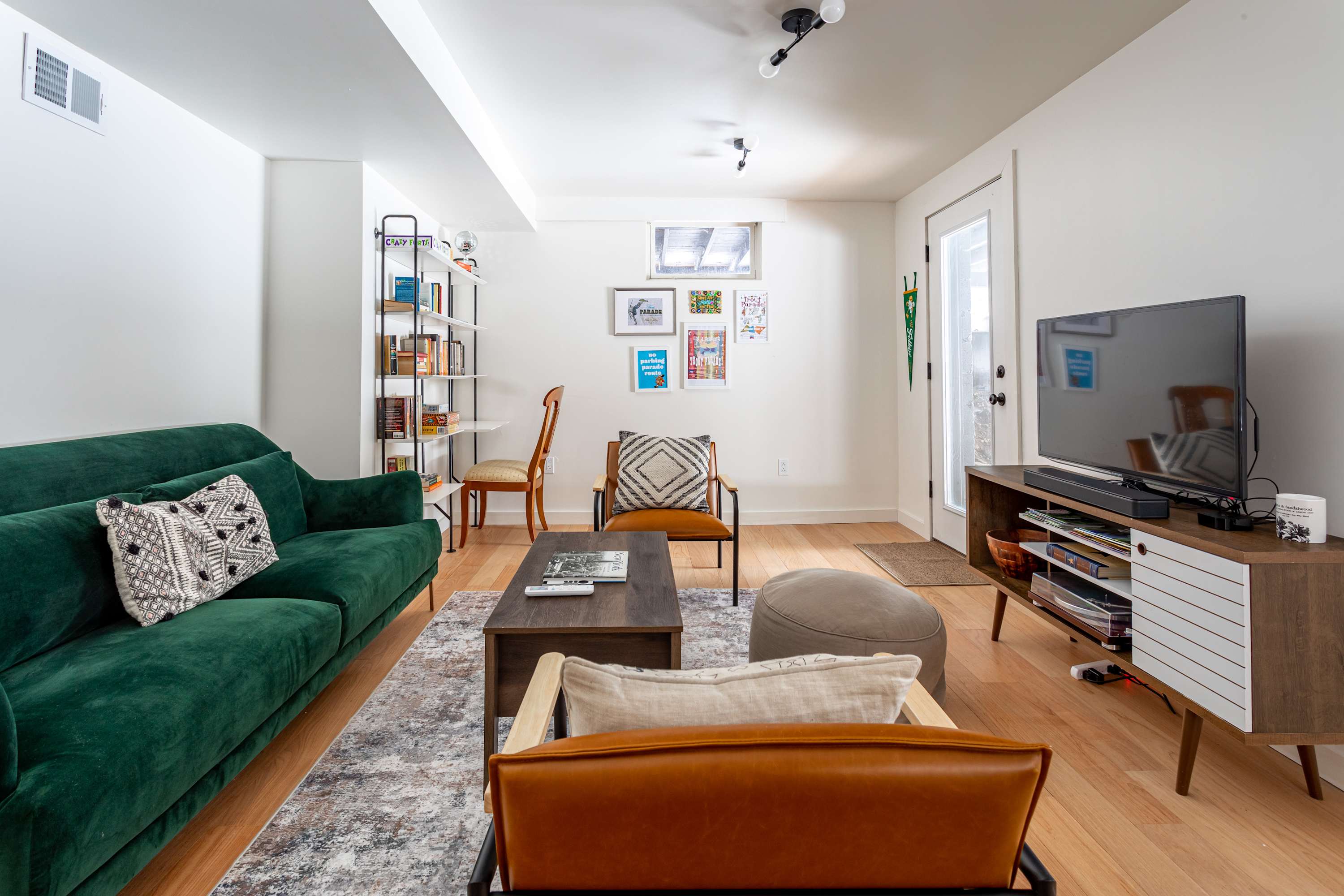 Bright basement living area with a green velvet sofa, a wooden coffee table, and a TV on a mid-century modern console. White walls and light wood floors create an airy feel, with a small window near the ceiling.