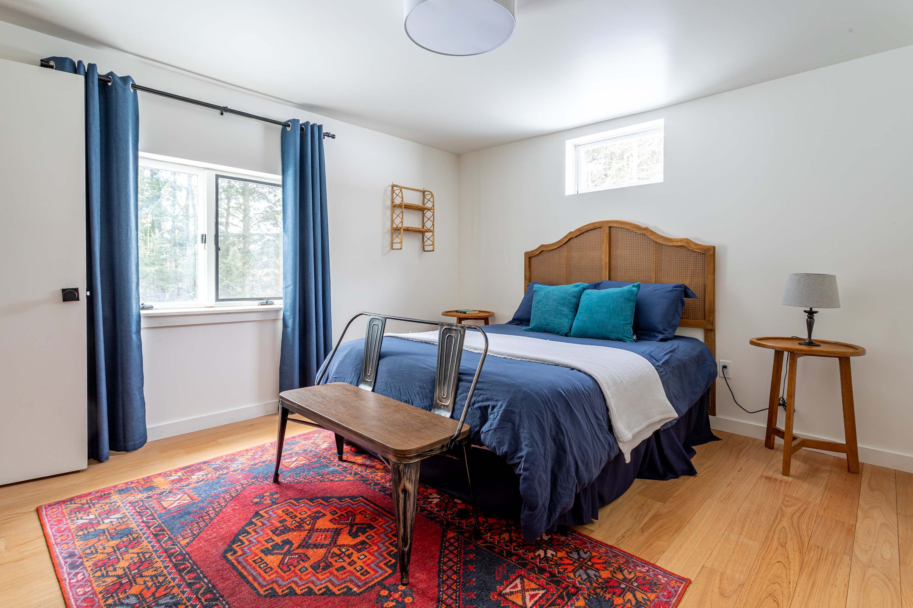 A cozy bedroom features a queen-sized bed with a rattan headboard, blue bedding, and a vibrant red patterned rug on light hardwood floors. A large window with blue curtains offers natural light.