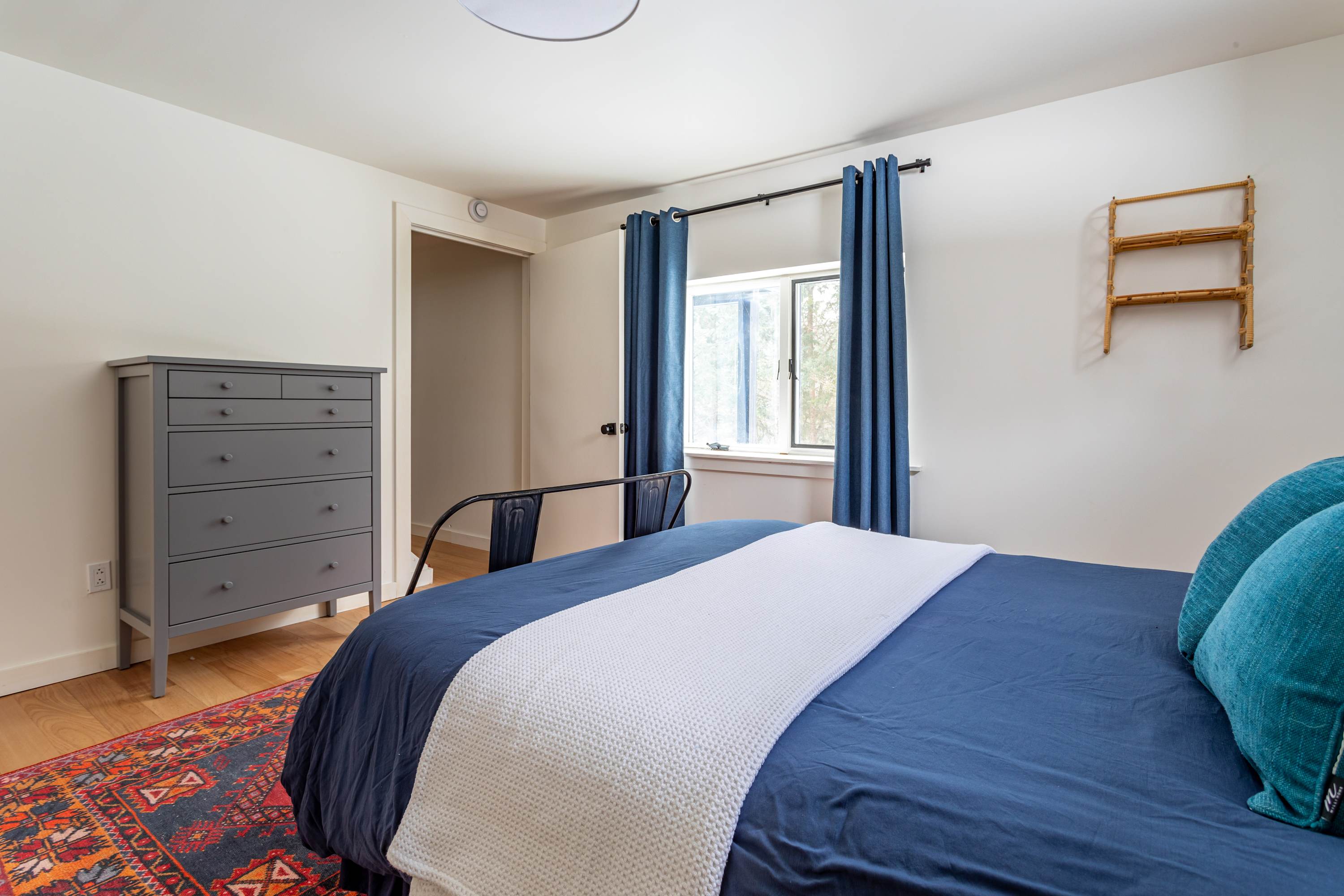 This bedroom view highlights a grey dresser with multiple drawers, positioned against a white wall, complementing the blue-themed bedding and red patterned rug on hardwood floors.