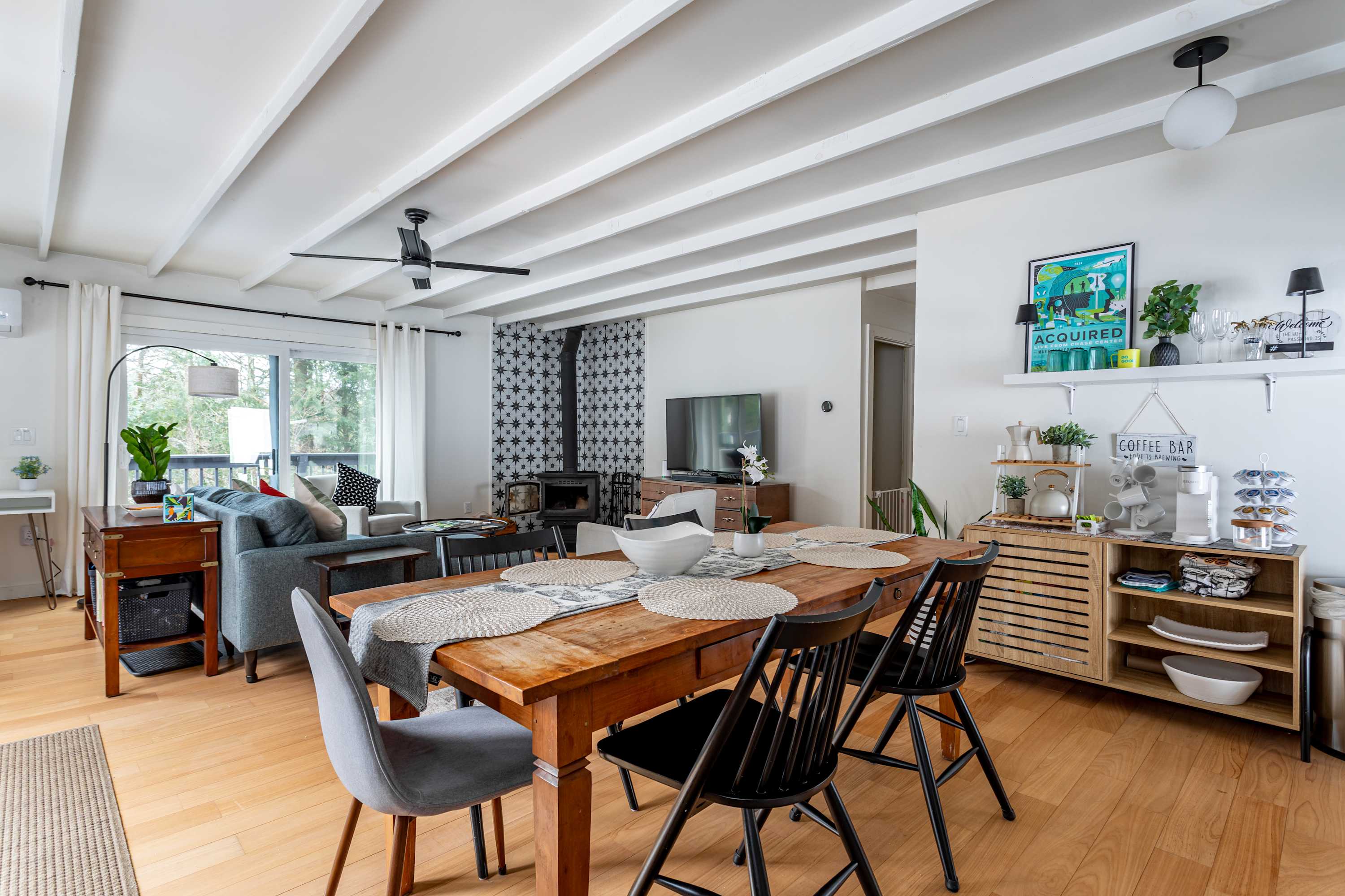 Open-concept dining and living area with a rustic wooden dining table, mixed seating, and a cozy living space featuring a woodstove against a patterned accent wall. White beamed ceilings add architectural interest.