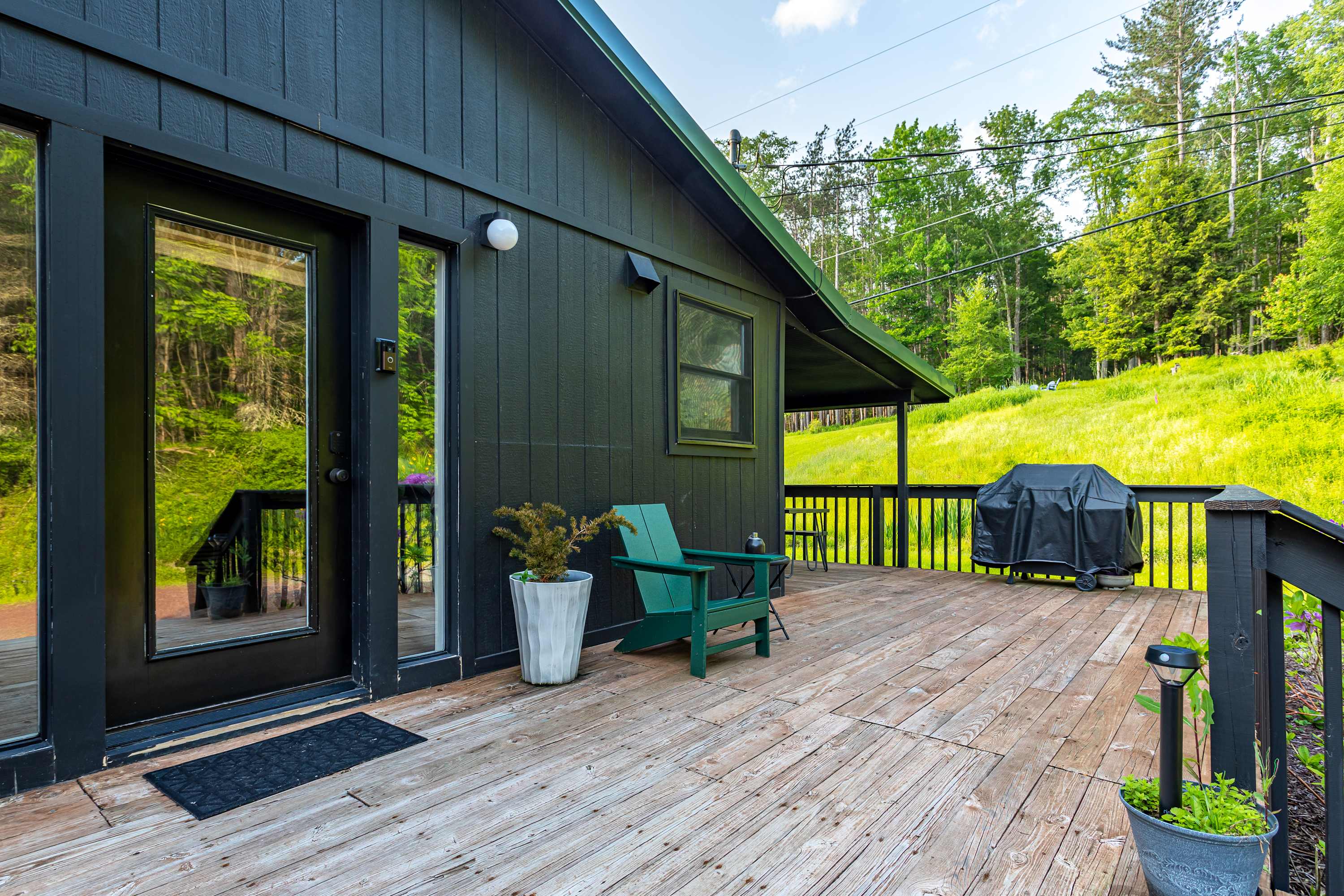 This deck view shows the black exterior of the house with a glass door, a green Adirondack chair, and a BBQ grill, set against a backdrop of green hills and trees, ideal for outdoor dining.
