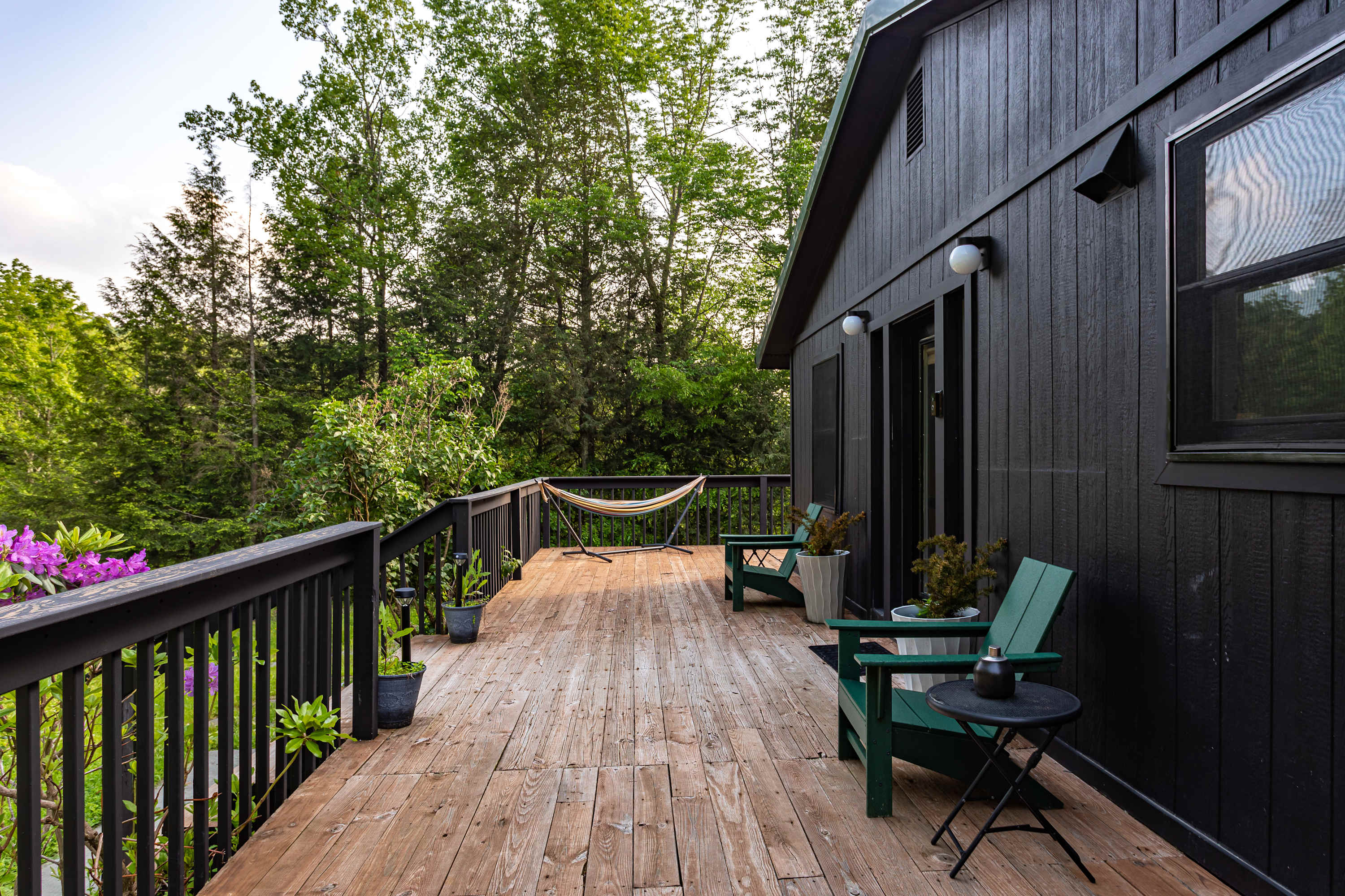 A spacious wooden deck extends into a lush green forest, featuring a striped hammock, green Adirondack chairs, and potted plants, perfect for outdoor relaxation and enjoying nature.