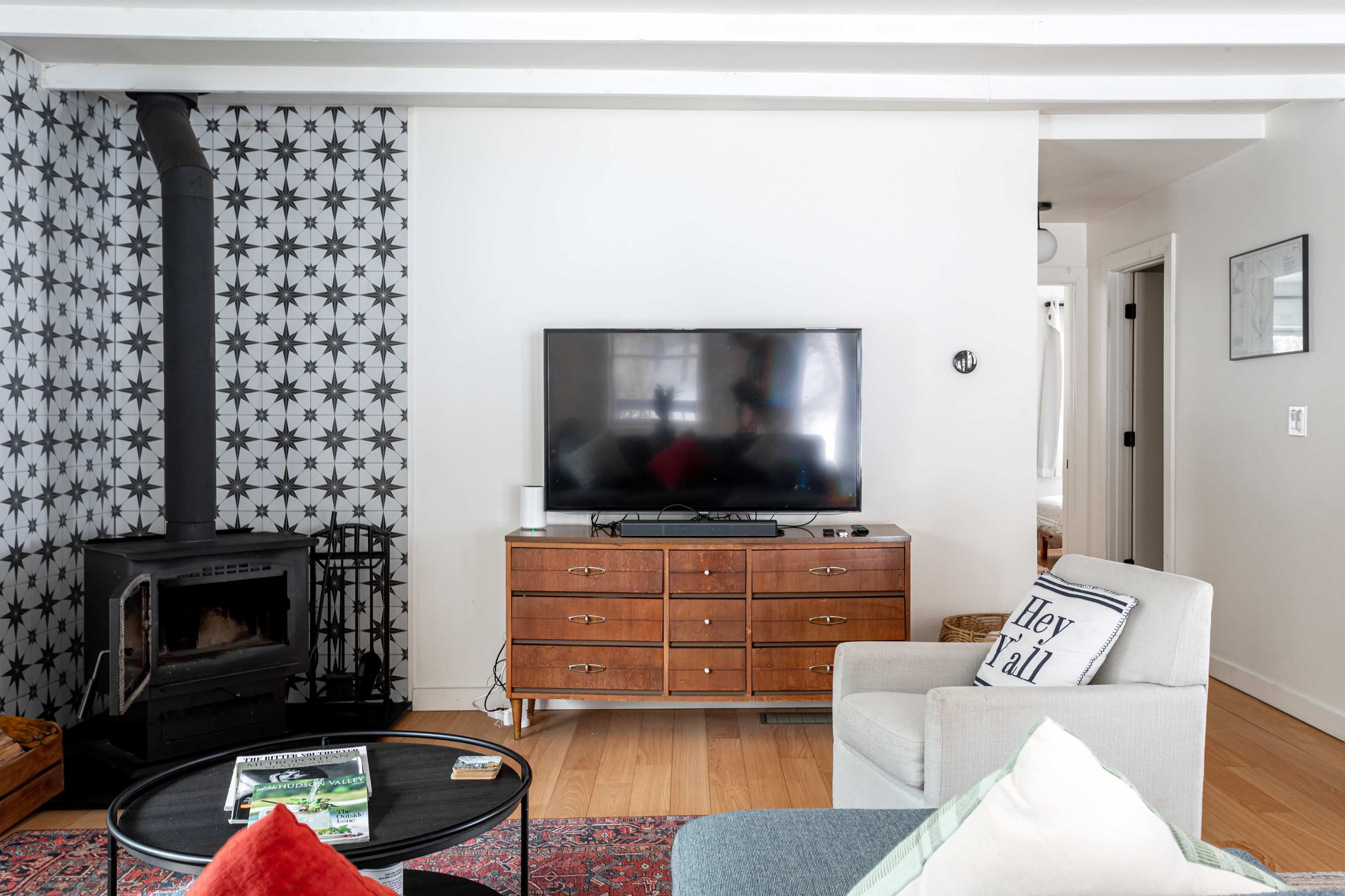 Detailed view of the living room's entertainment area, featuring a large flat-screen TV on a mid-century modern wooden dresser, adjacent to a woodstove with a star-patterned tile backdrop. A comfortable armchair is also visible.