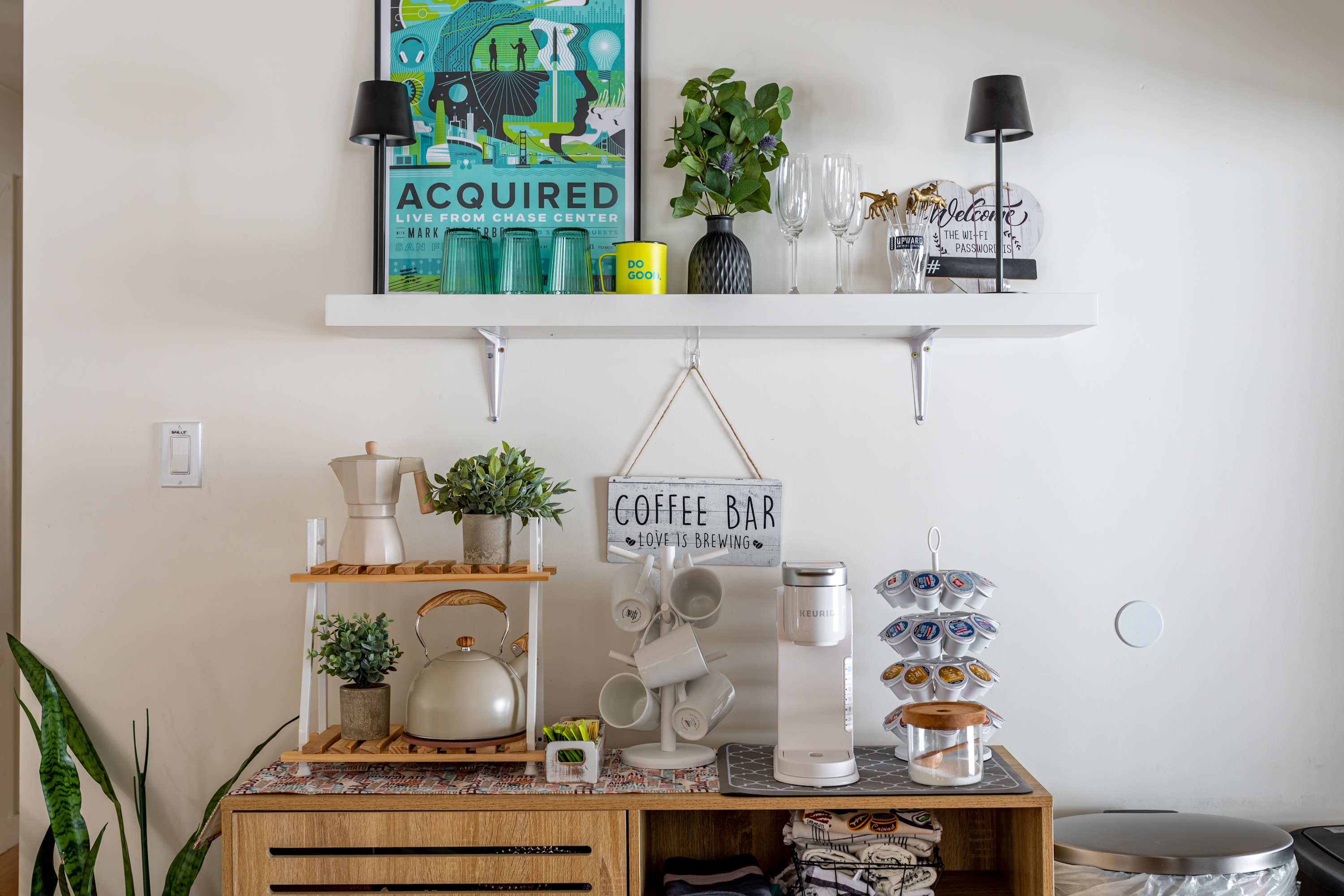 A dedicated coffee bar nook featuring a wooden cabinet, a two-tiered shelf with coffee-making appliances, and a wall-mounted "Coffee Bar" sign. Various mugs and coffee pods are neatly organized.