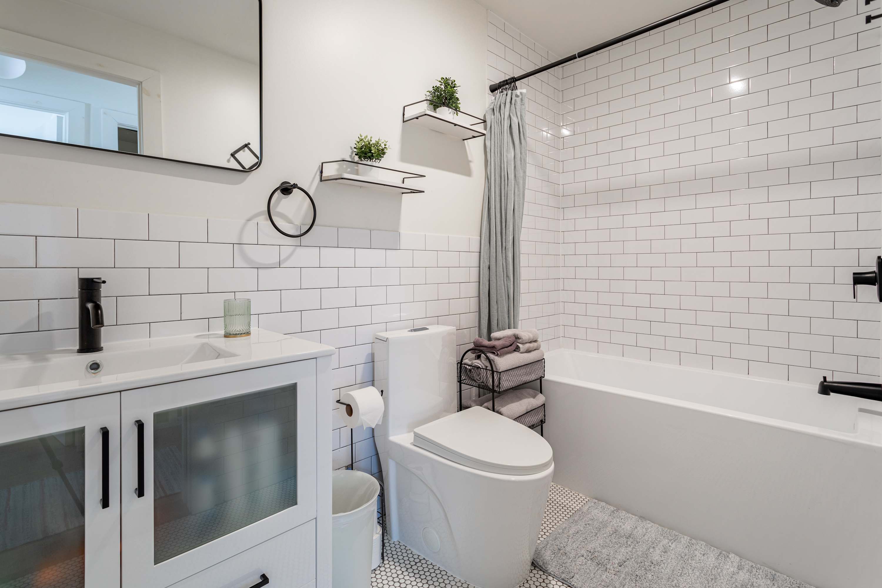 Modern bathroom with white subway tile walls, a white vanity with a black faucet, a toilet, and a bathtub with a shower. Hexagonal floor tiles add a touch of pattern.