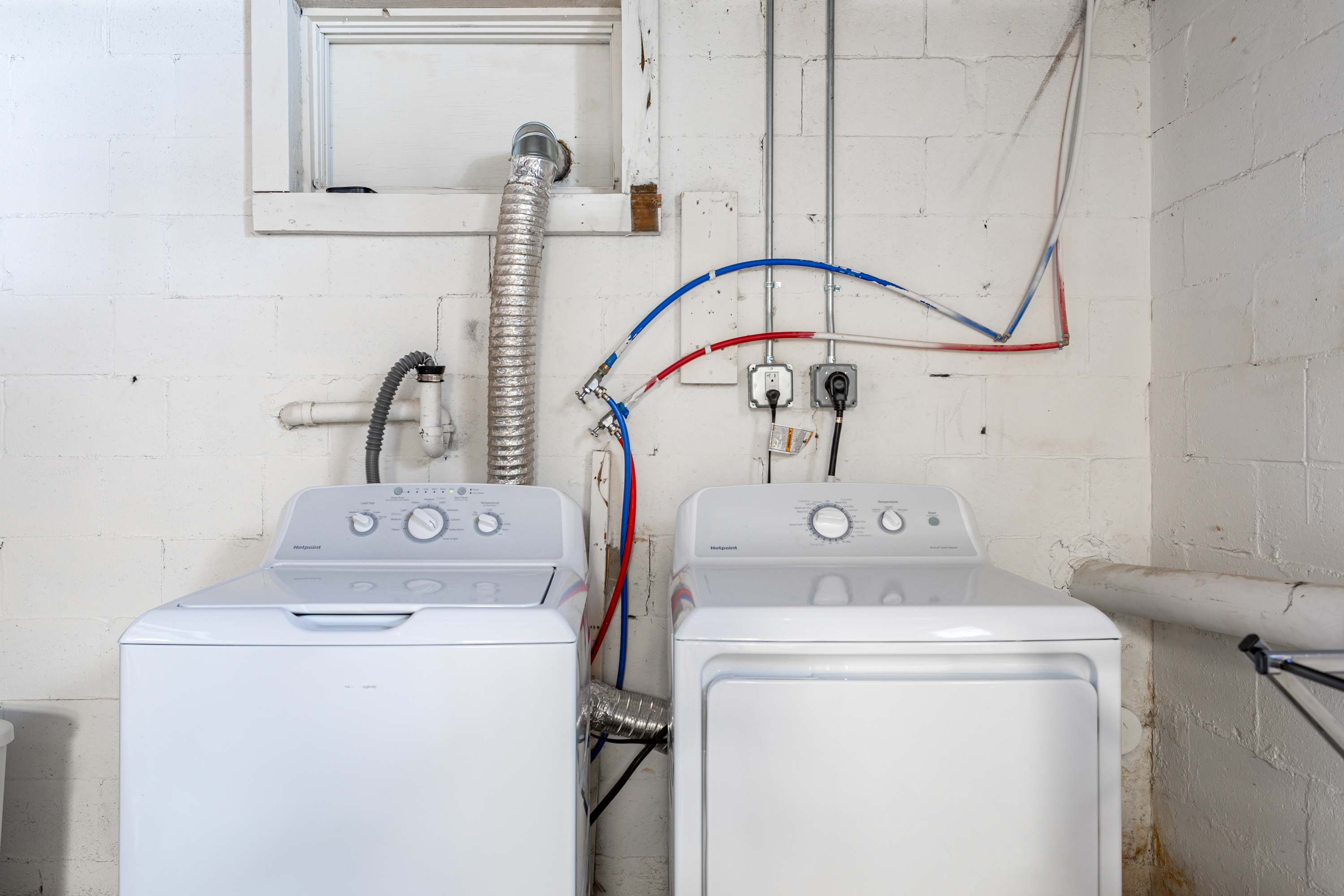 A functional laundry area features a white top-loading washing machine and a matching dryer, with exposed plumbing and ductwork against a white block wall, indicating utility space.