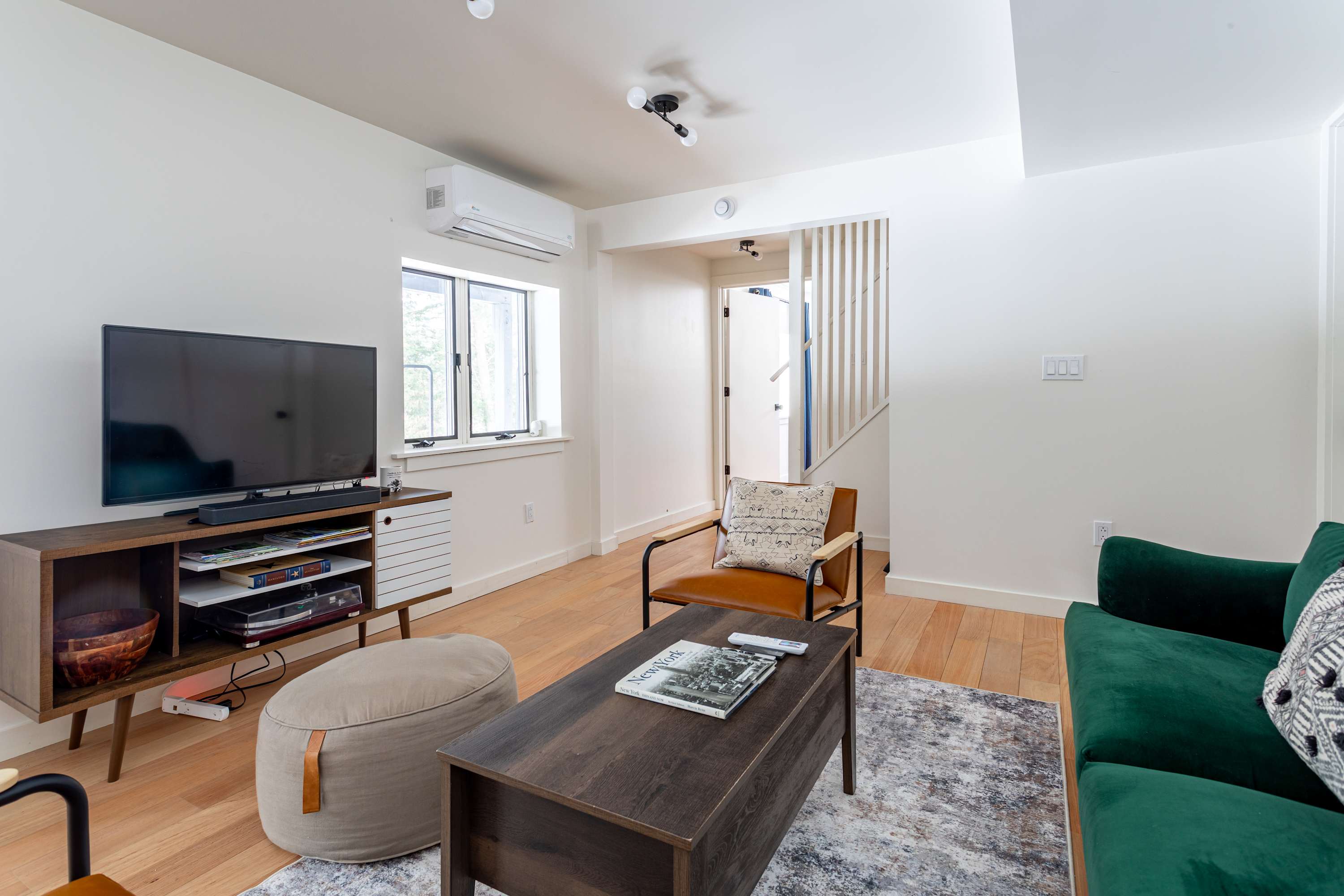 This living room view shows a flat-screen TV on a wooden media console, a window with a mini-split AC unit above it, and a comfortable armchair with a pouf ottoman, all on light hardwood floors.