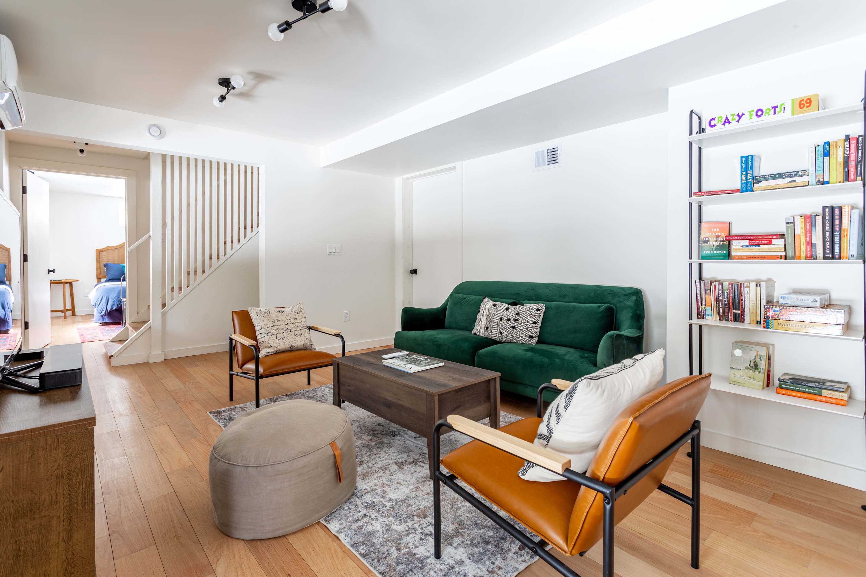 A modern living room features a dark green sofa, two tan leather armchairs, and a dark wood coffee table on a patterned rug. A white bookshelf filled with books stands against the wall, offering entertainment.