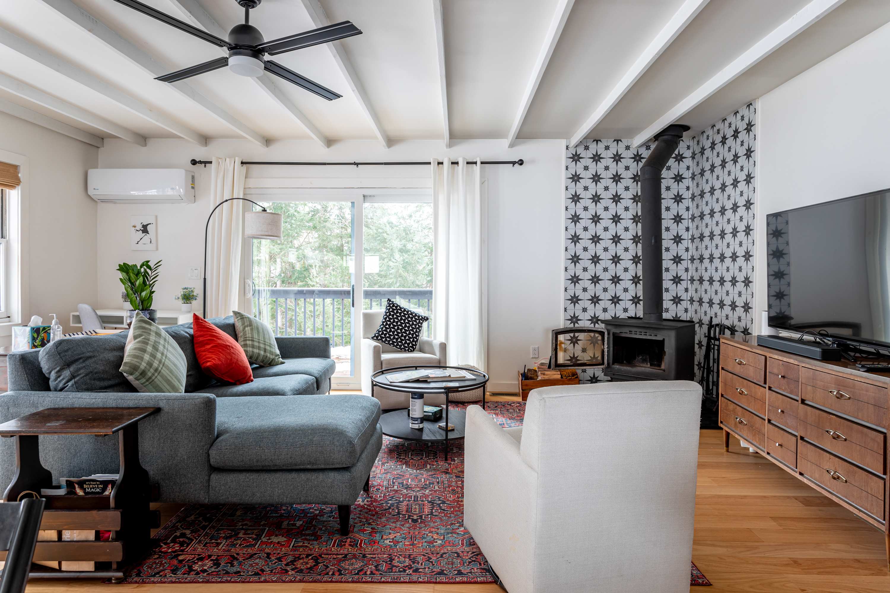Inviting living room with a grey sectional sofa, a round coffee table, and a prominent woodstove set against a striking black and white patterned accent wall. A large flat-screen TV is mounted on a wooden console.
