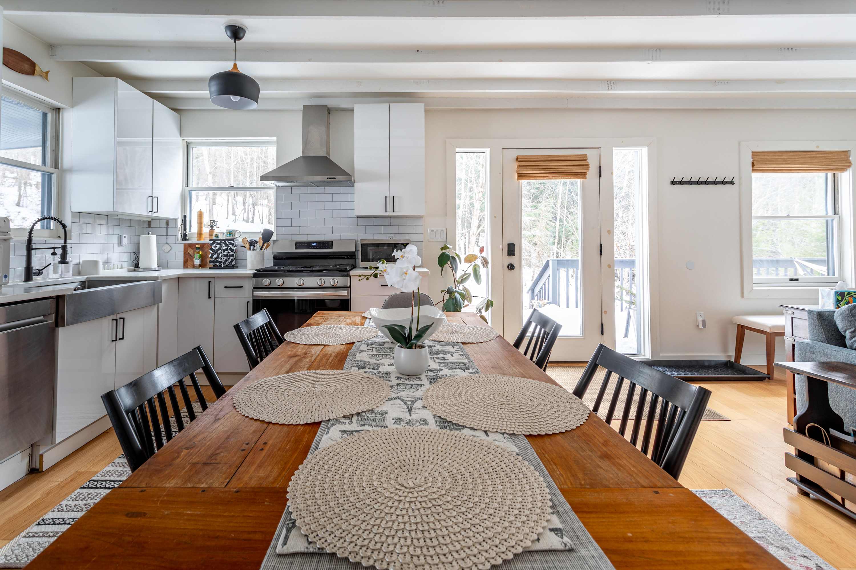 Bright kitchen and dining area with white cabinetry, subway tile backsplash, stainless steel appliances, and a large wooden dining table. A glass-paned door leads to an outdoor deck.