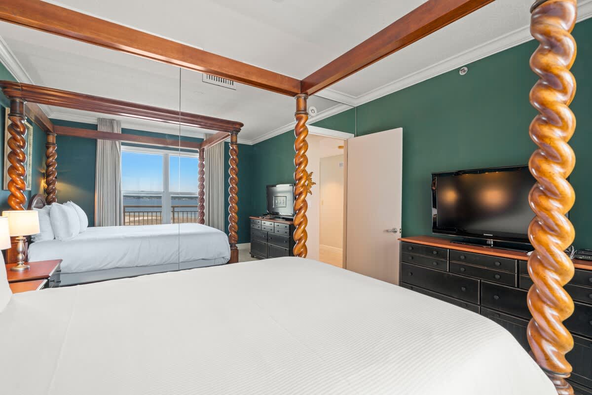 A spacious bedroom featuring two four-poster beds with white bedding, dark green walls, and a large window offering panoramic ocean views. A flat-screen TV sits on a dark dresser.