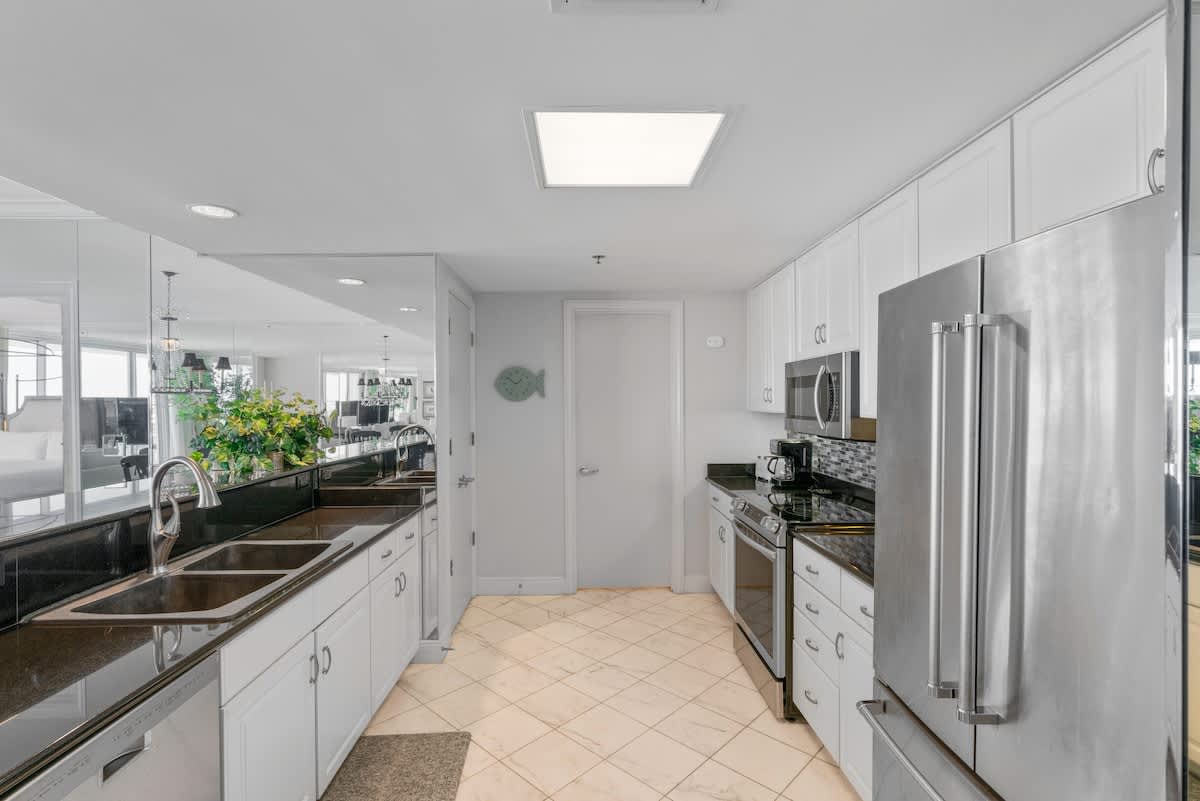 A galley kitchen features white cabinetry, black granite countertops, and stainless steel appliances, with a double sink on the left and a large mirror reflecting the dining area. The floor is tiled in a diagonal pattern.