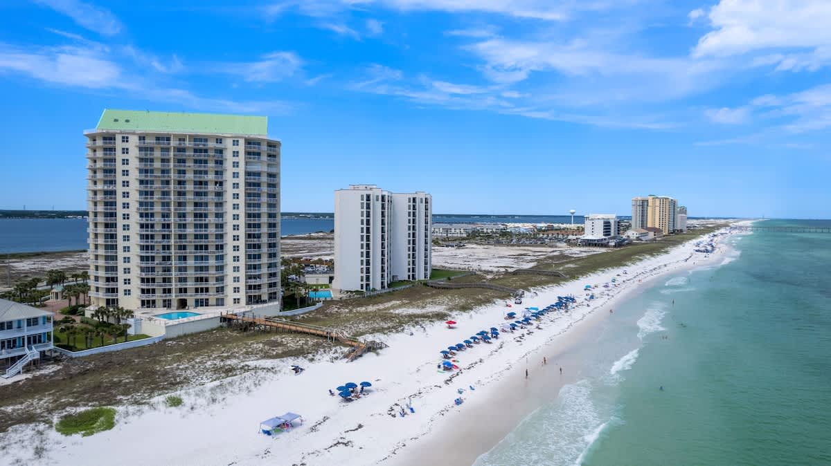 Expansive aerial view showcasing multiple tall condominium buildings along a vast white sand beach, with the turquoise ocean stretching to the horizon. The property features a pool, sports courts, and a boardwalk, all under a bright blue sky.