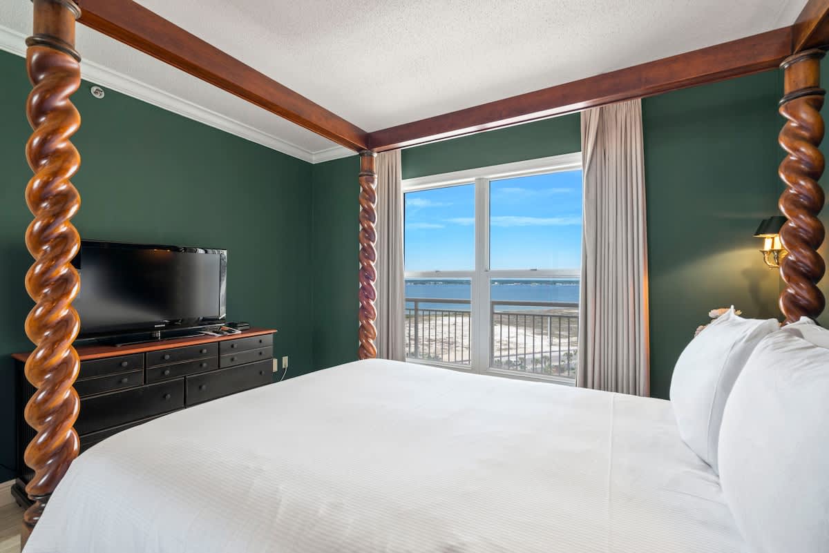 A cozy bedroom with a four-poster bed, white bedding, and dark green walls. A large window provides a stunning view of the ocean and coastline. A flat-screen TV is on a dark dresser.