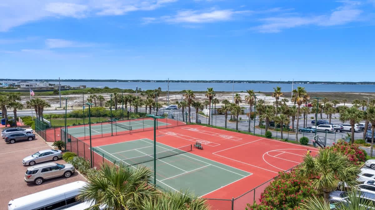 Elevated view of red and green multi-sport courts, including two tennis courts, a basketball court, and shuffleboard areas, bordered by a parking lot and palm trees. A wide expanse of calm blue water and a distant shoreline are visible under a clear sky.
