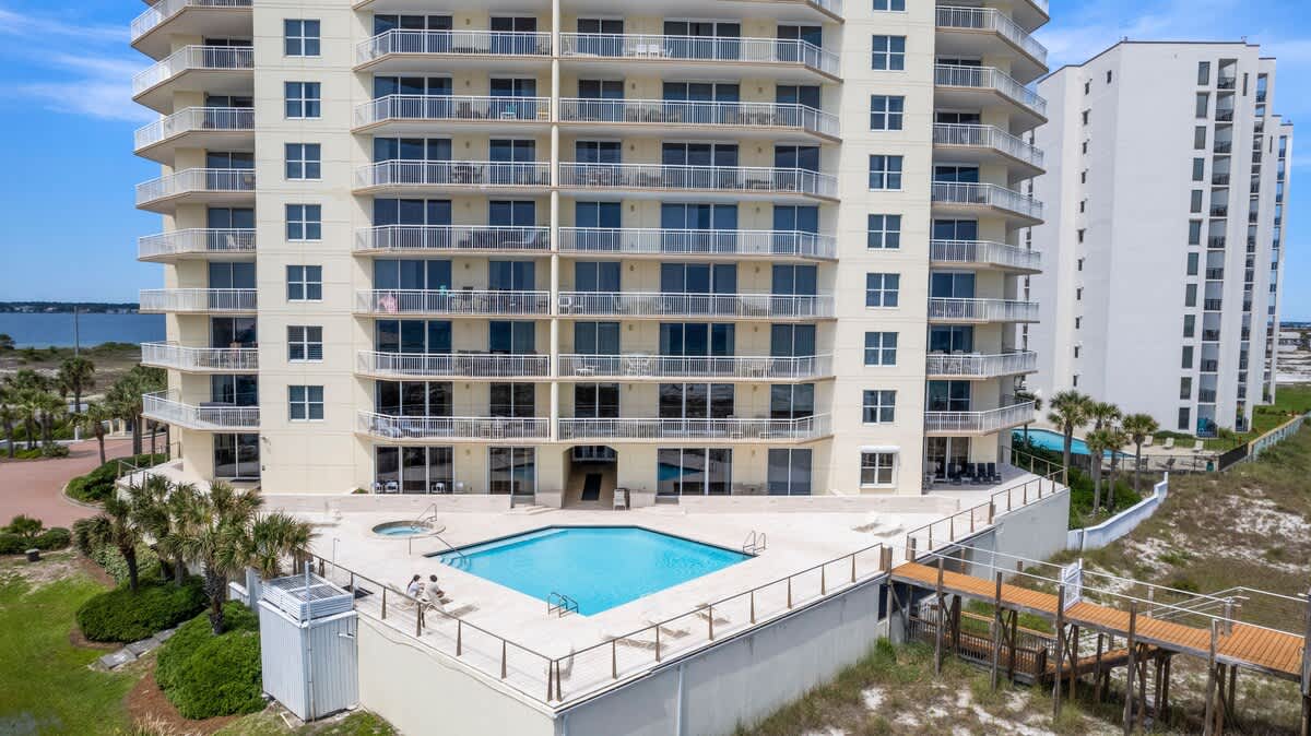 Aerial view of a tall, light-colored condominium building with multiple balconies, a rectangular outdoor swimming pool with a hot tub, and a wooden boardwalk extending towards a distant body of water. Palm trees and sandy dunes are visible around the property.