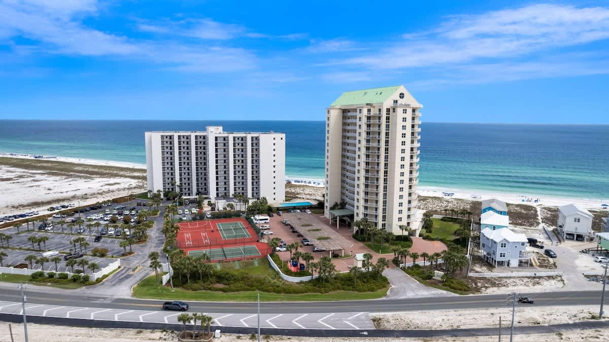 High aerial view of two large, light-colored condominium buildings, a spacious parking area, and red multi-sport courts, all set against the backdrop of a wide, calm ocean and a clear blue sky. A road runs in front of the property.