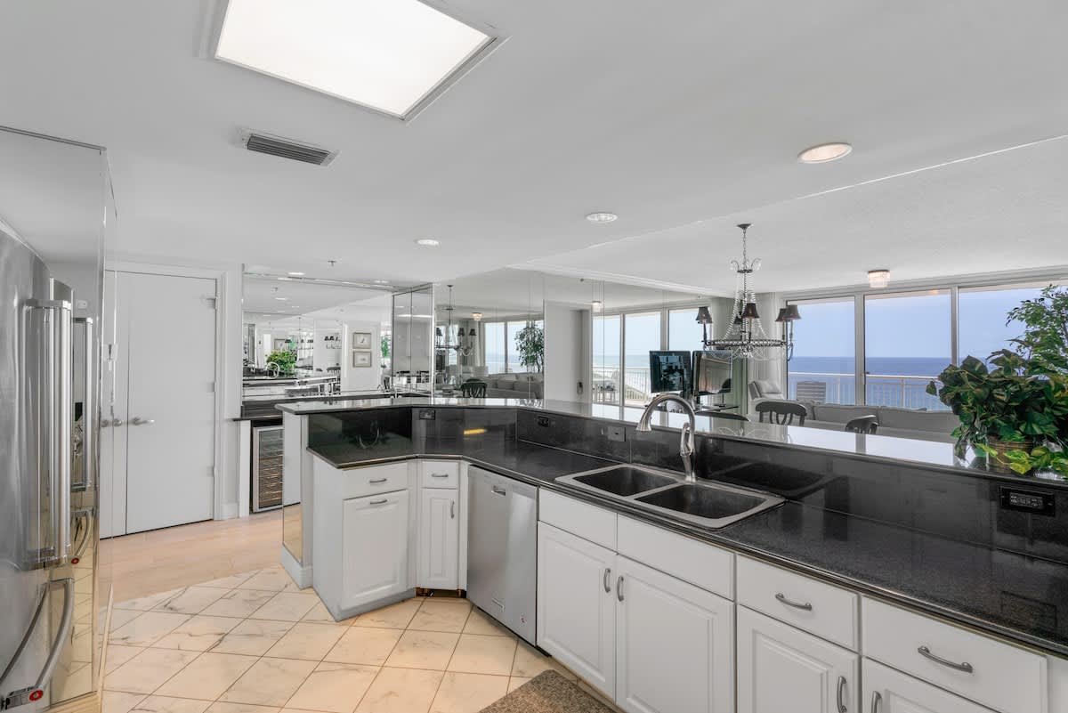 A close-up of the kitchen area, highlighting the black granite countertops, double sink, and white cabinetry. Mirrored surfaces on the island and walls reflect the bright, open space.