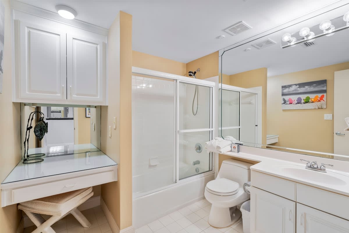 A bright bathroom with light beige walls, a white vanity with a single sink, a toilet, and a shower/tub combo with a sliding glass door. A large mirror spans the wall above the vanity.