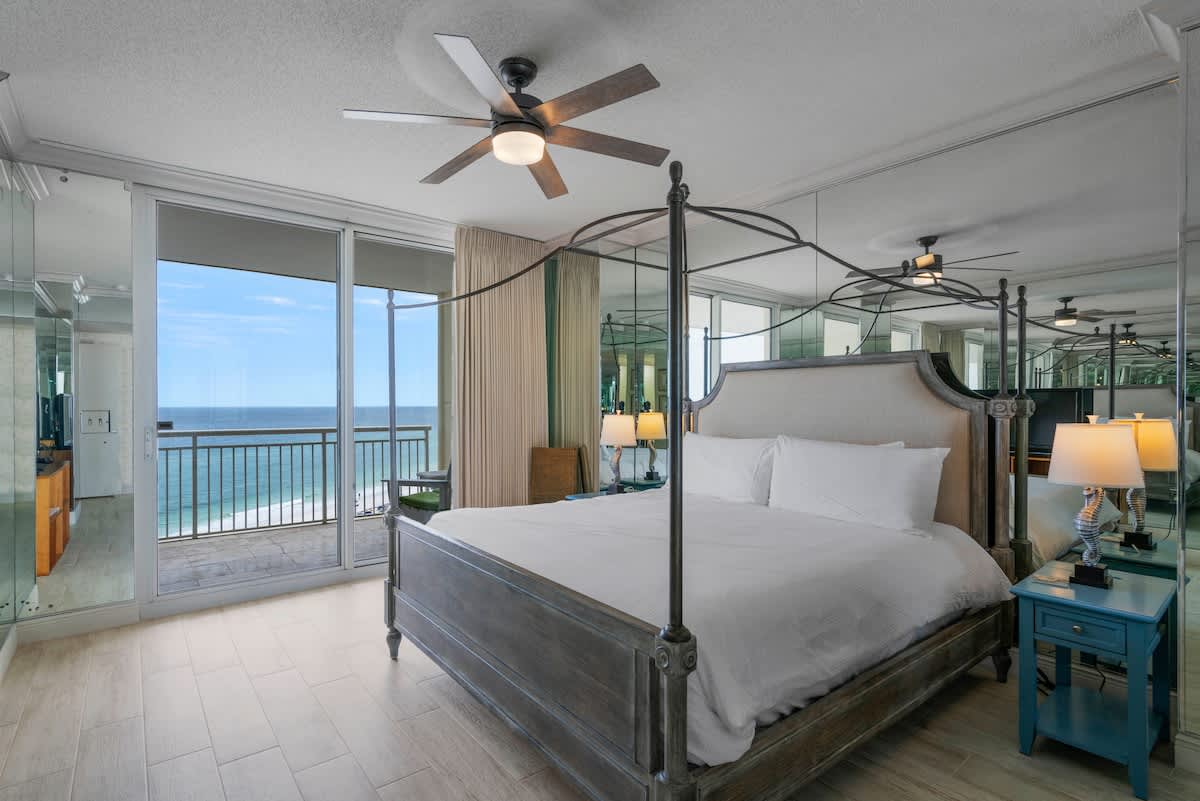 A bright bedroom features a light gray upholstered canopy bed with white bedding, mirrored walls, and a ceiling fan. Sliding glass doors open to a balcony with stunning ocean views.