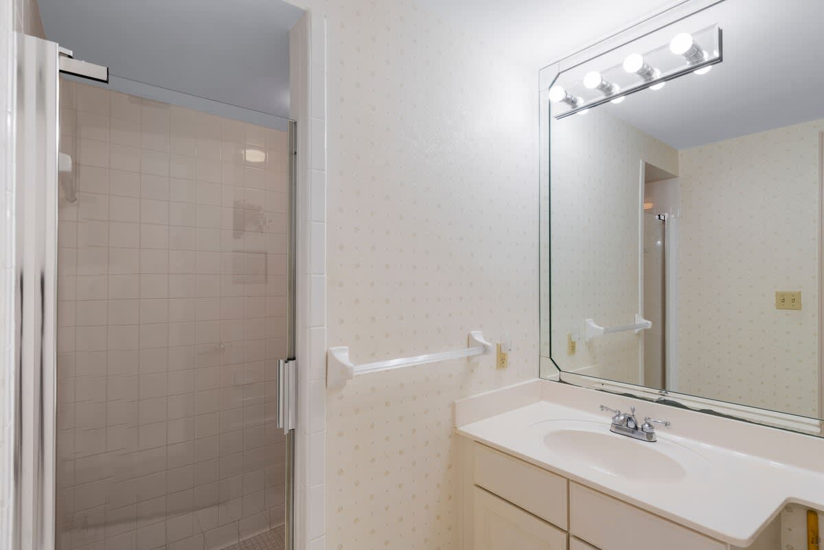 A bathroom with light patterned wallpaper features a single vanity with a large mirror and a separate walk-in shower with white tiles. The vanity has a light fixture above the mirror.