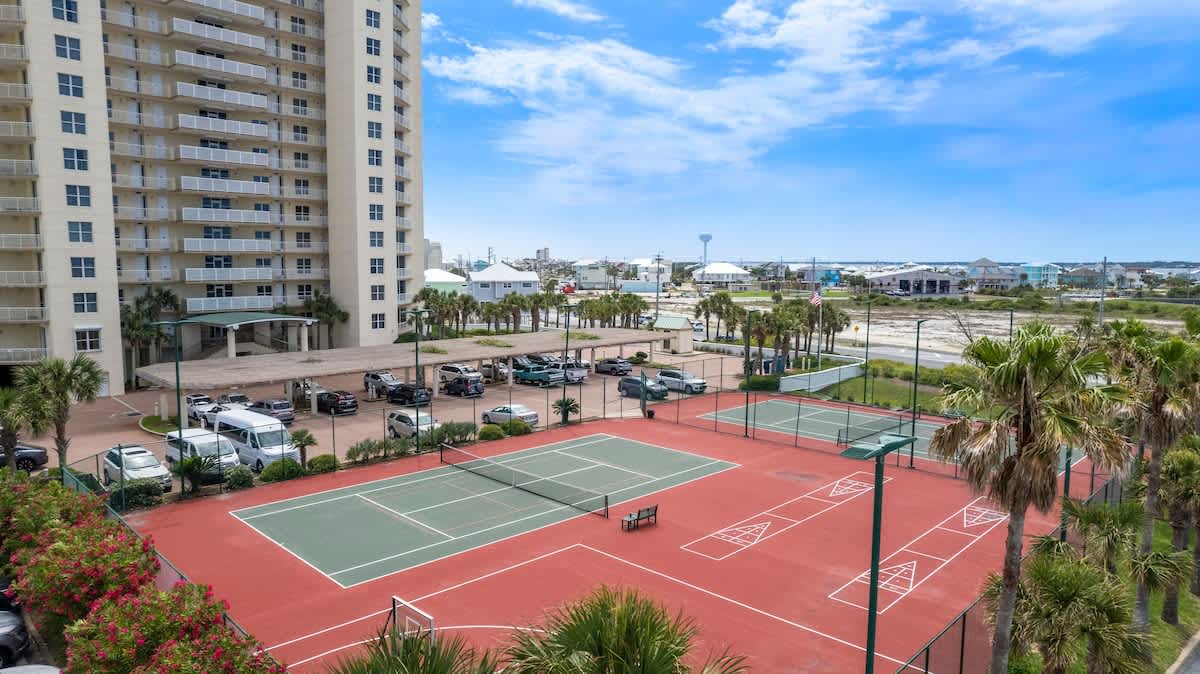 Aerial view of a large condominium building overlooking a parking lot filled with cars, and a multi-sport court area featuring two tennis courts, a basketball hoop, and shuffleboard courts, all surrounded by palm trees. A distant town and water tower are visible in the background.