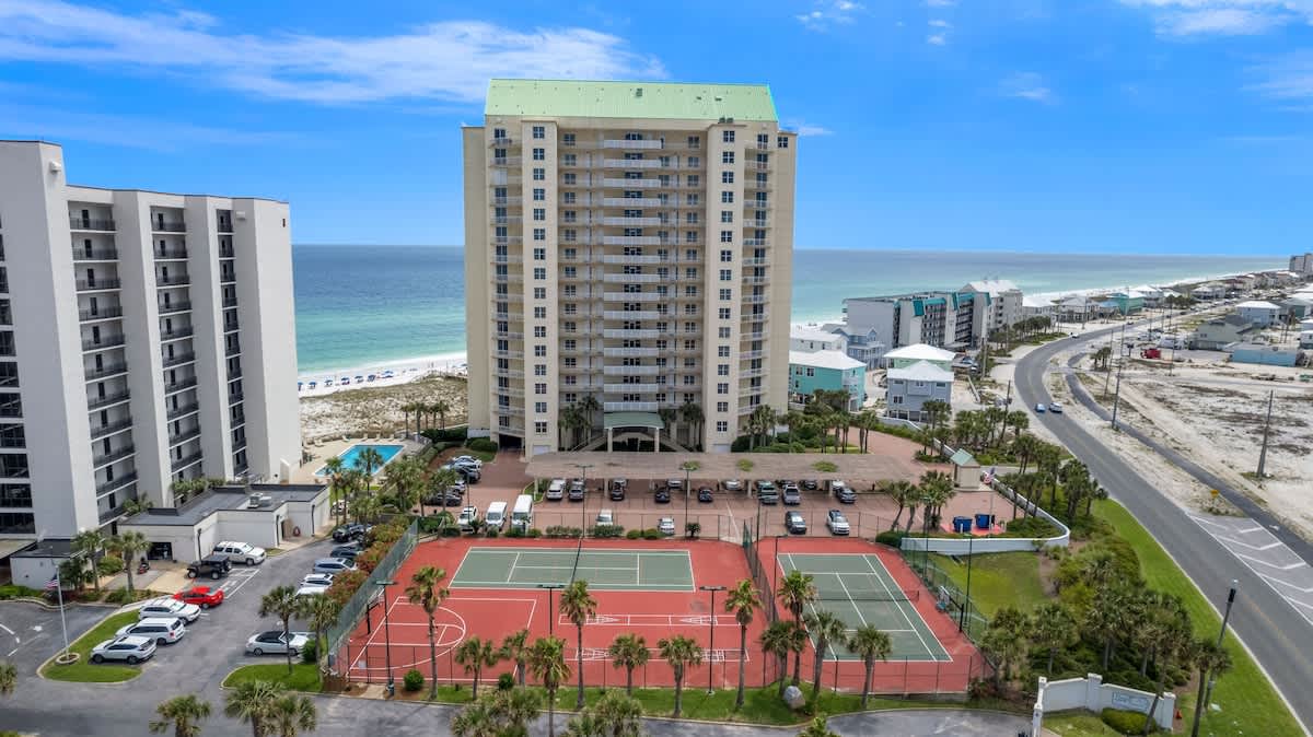 Wide aerial shot showcasing two large condominium buildings, extensive parking areas, and red multi-sport courts with tennis and basketball. The property is situated directly on a white sand beach with turquoise ocean waters under a bright blue sky.