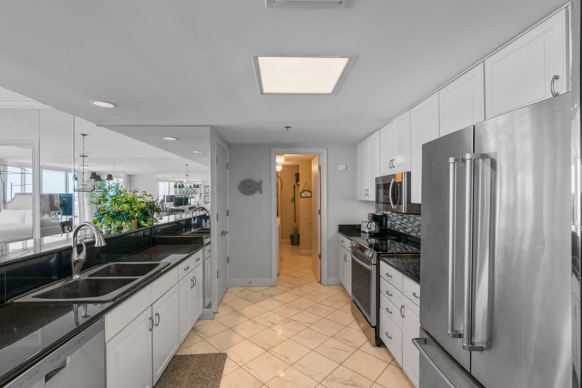 A bright galley kitchen with white cabinets, black granite countertops, and stainless steel appliances, featuring a double sink and a mirrored wall reflecting the adjacent dining space. A doorway at the end leads to another room.